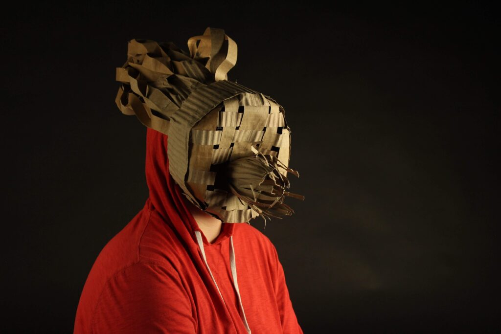 A figure sits in front of a black backdrop, wearing an orange hoodie. on their head is a mask made of cardboard abstracting their face. The surface is woven and curls and obstructs. 